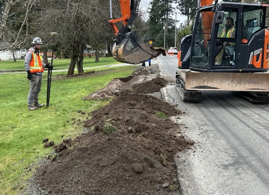 Photo of an excavator digging a trench along the side of a road as part of the sidewalk project