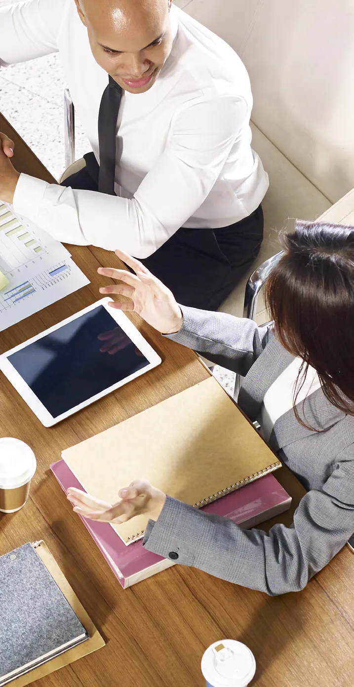 Two people in business attire sitting at a table discussing documents and a tablet.