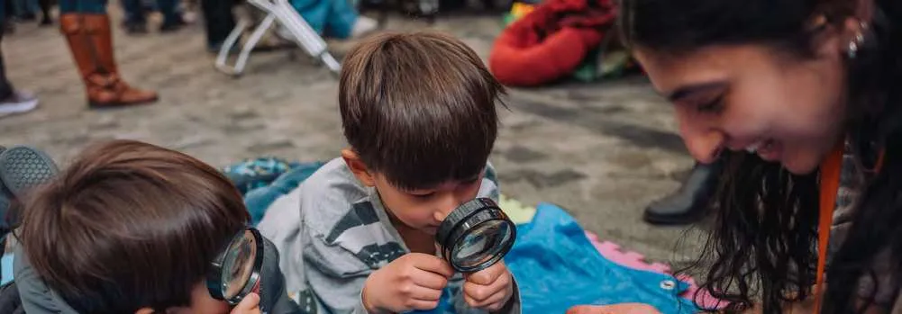 Two young children using magnifying glasses to look at soil held by an instructor