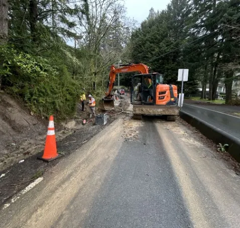 Construction scene on Atkins Road with an excavator and workers preparing the sidewalk base