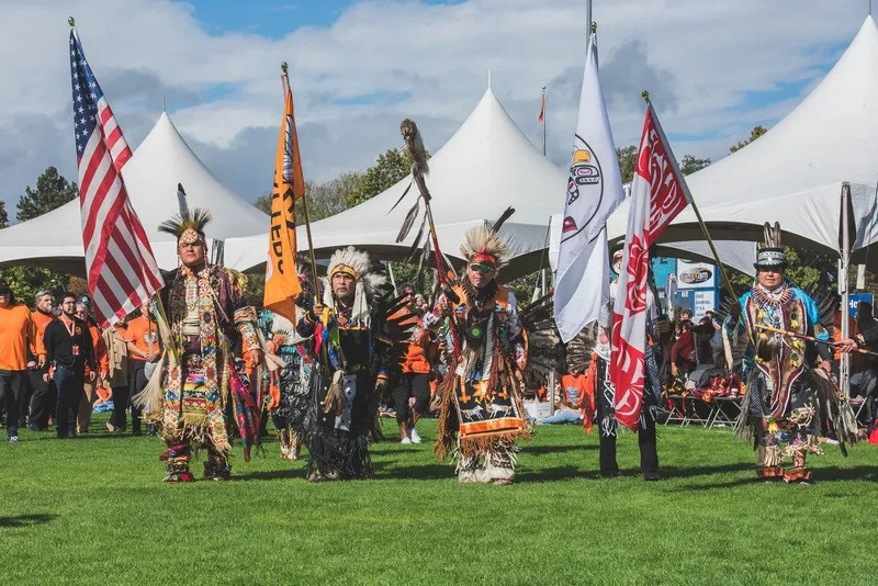 Powwow participants in traditional regalia carrying various flags across a grass field