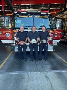 Three firefighters in uniform standing in a fire hall bay