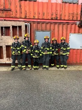 Six new fire recruits in full firefighting gear posing in front of training containers
