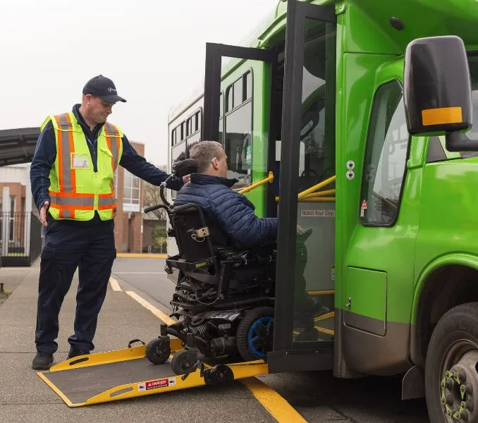 A handyDART driver assisting a passenger in a power wheelchair onto a bus using an automated ramp
