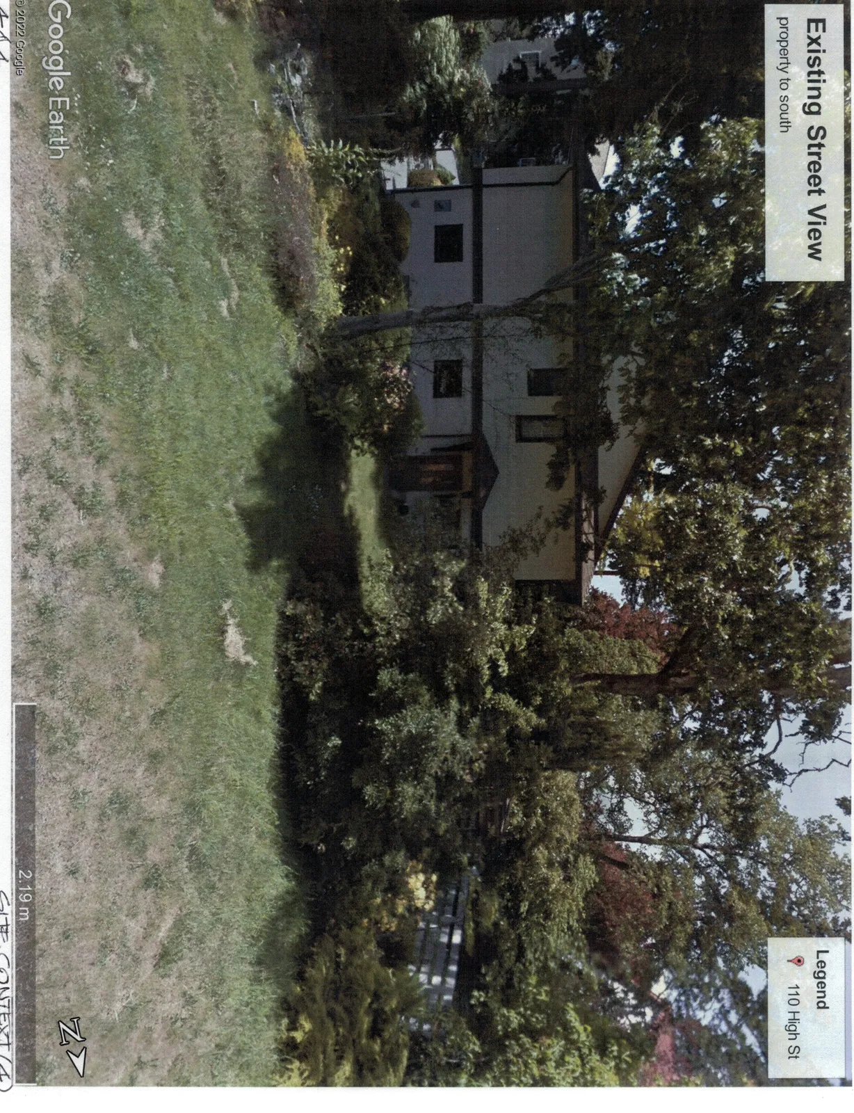 Street view photograph of the neighboring property to the south, showing a two-story residence with light-colored siding and dark trim, partially obscured by mature trees and shrubs.