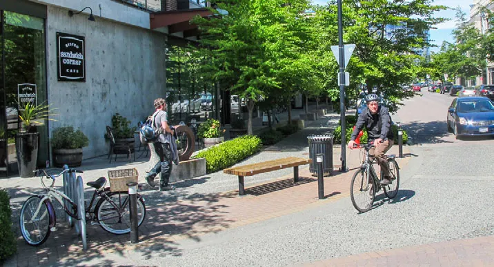 Urban streetscape with a person walking, a person on a bicycle, and a bike rack near a cafe