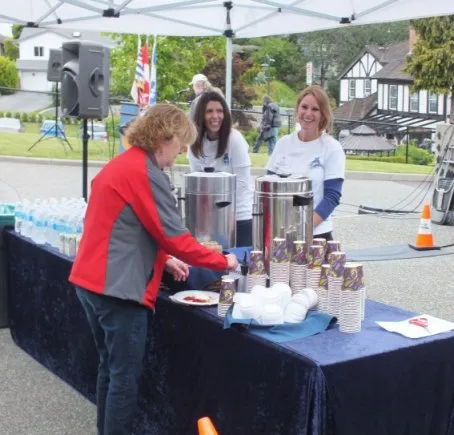 Three women standing behind a table serving food and drinks under an outdoor canopy.