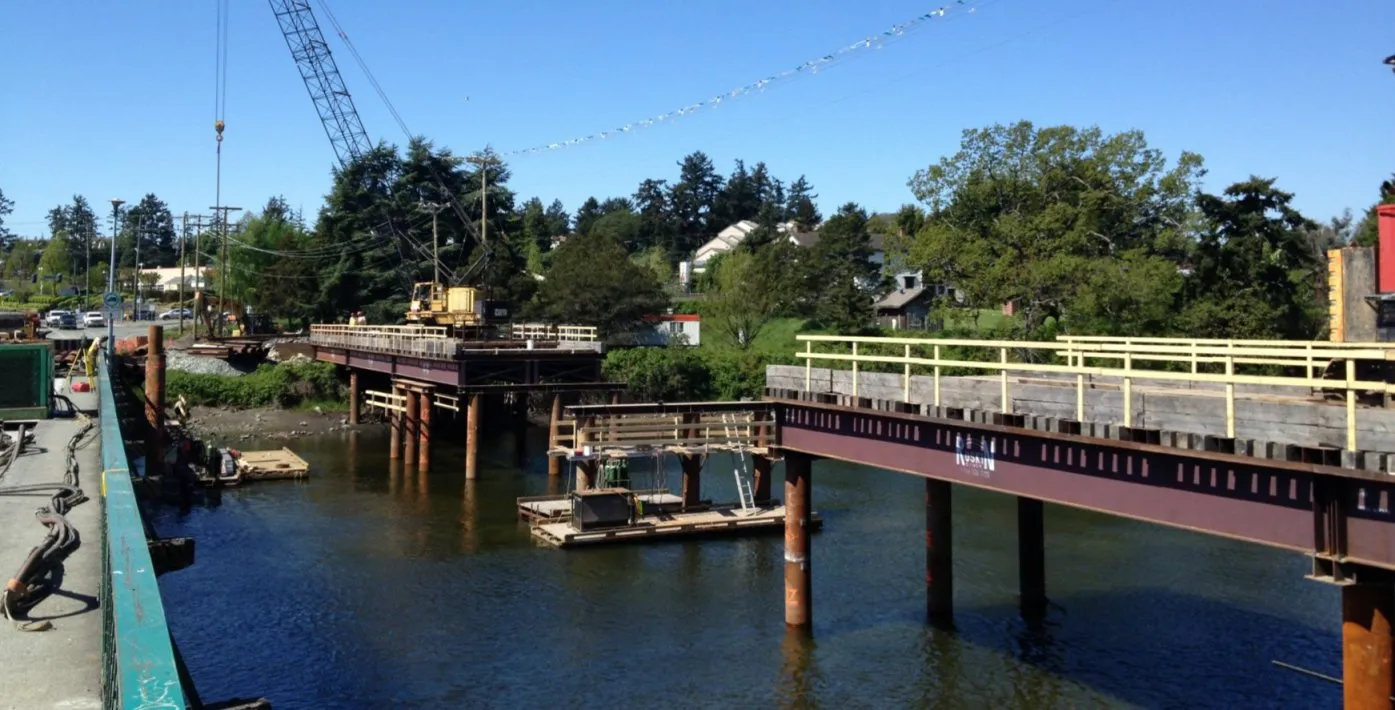 A view of water with a railway track in the foreground and forested hills across the inlet.