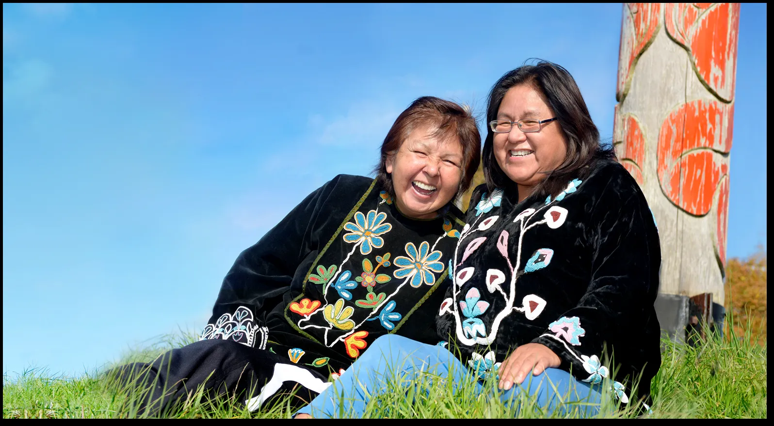 Two women laughing together while sitting on the grass near a totem pole