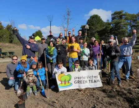 A large group of community volunteers holding gardening tools and a sign in a coastal park setting