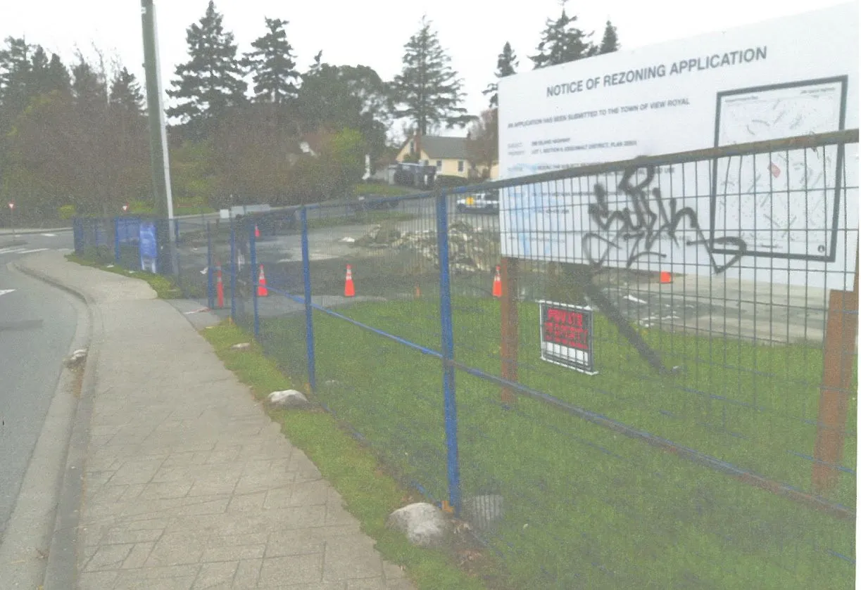 Photograph of the existing construction site featuring a blue metal fence and a "Notice of Rezoning Application" sign