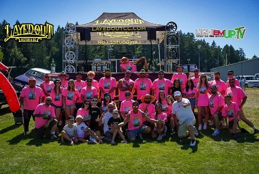 A large group of people wearing pink shirts posing for a photo in front of a stage at the Layedout Field of Dreams Car Show