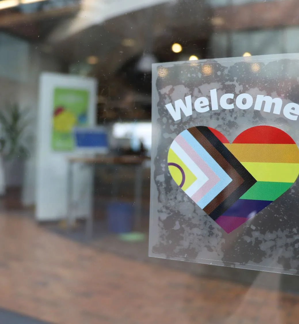 Close-up of a 'Welcome' sign with a pride progress heart on a glass door.
