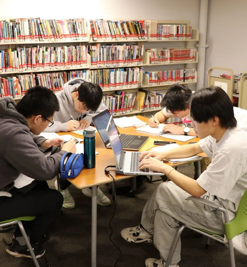 Four students sitting at a table in a library, working on laptops and studying.