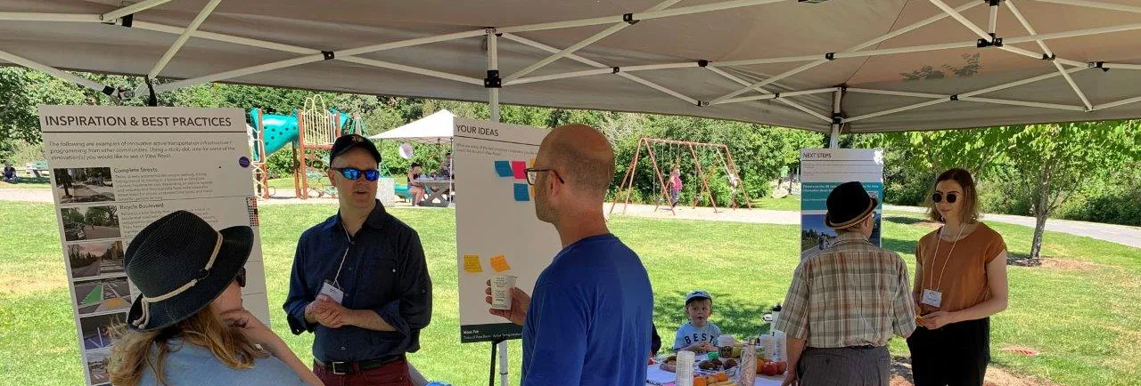 People gathered under a canopy at an outdoor public event, looking at display boards titled "Inspiration & Best Practices" and "Your Ideas".