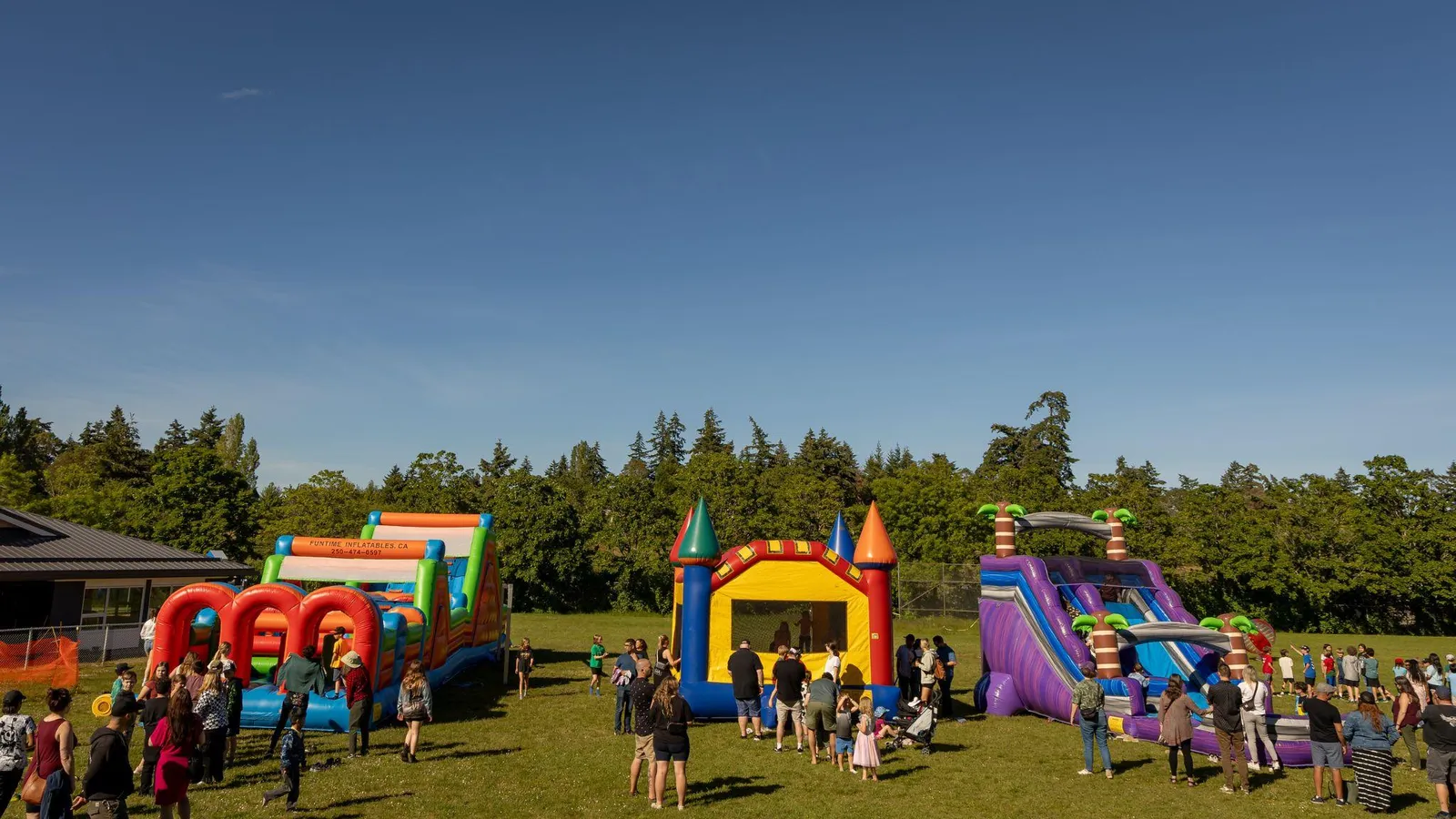 A photo showing families and children at a fair with several large, colorful inflatable structures, including a bouncy castle and slides, set up on a grassy field with a line of trees in the background.