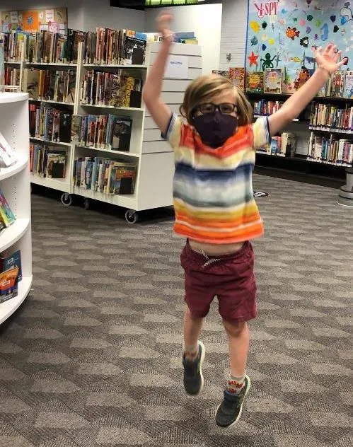 A young child wearing a face mask jumping excitedly in a library book aisle.
