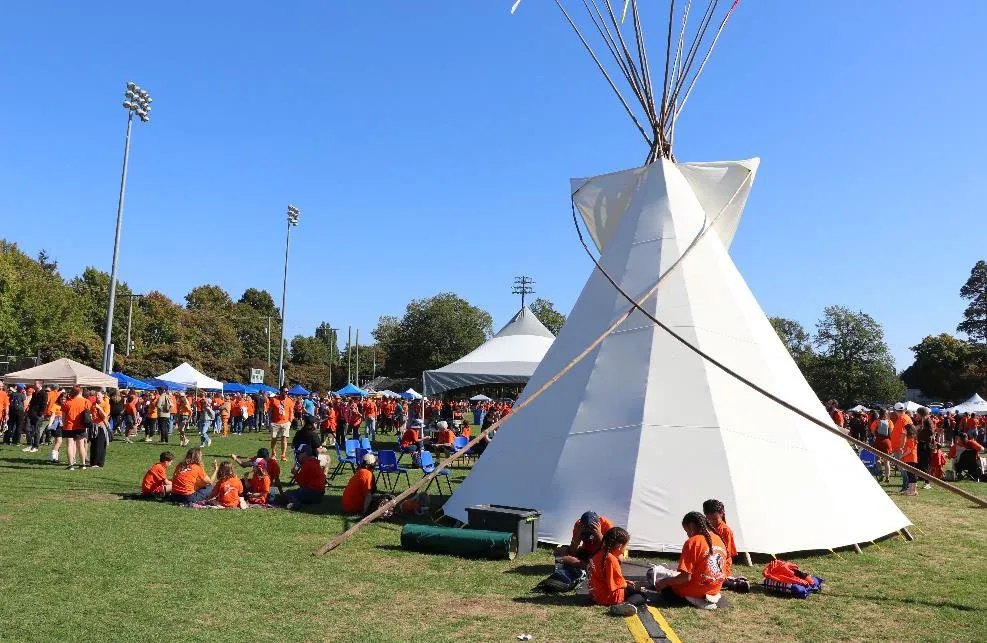 A large white tipi set up on a grassy field during an outdoor community event with many attendees wearing orange shirts.