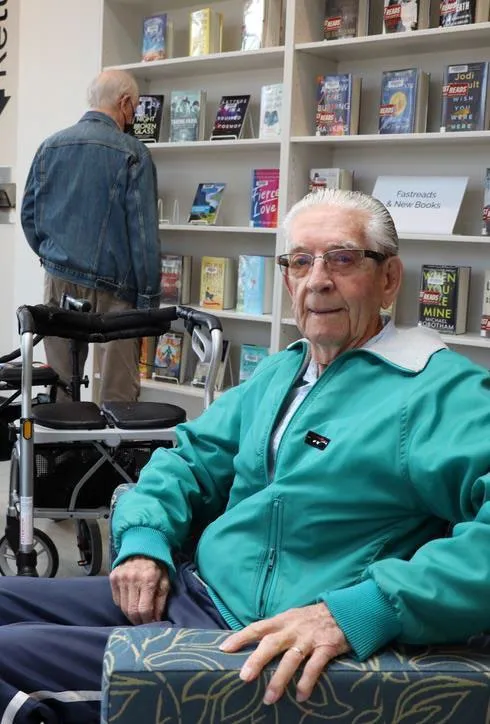 An elderly man smiling in a library setting, with bookshelves and another patron visible in the background.