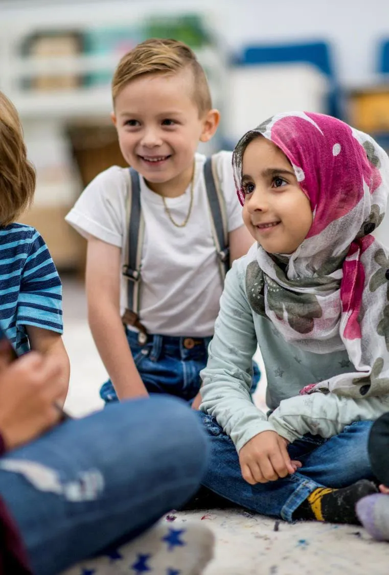 A group of young children of diverse backgrounds sitting together on the floor and smiling.