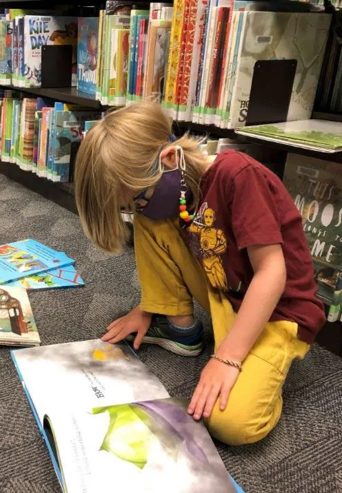 A young child sitting on a library floor intently reading an open picture book.