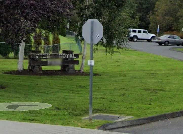 A photo of a sign post with the sign turned or missing in a grassy area, with a wooden "VIEW ROYAL" monument in the background.