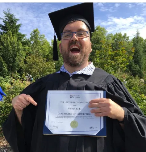 Nathan Bodie in a graduation cap and gown, holding his certificate of completion from the University of Victoria