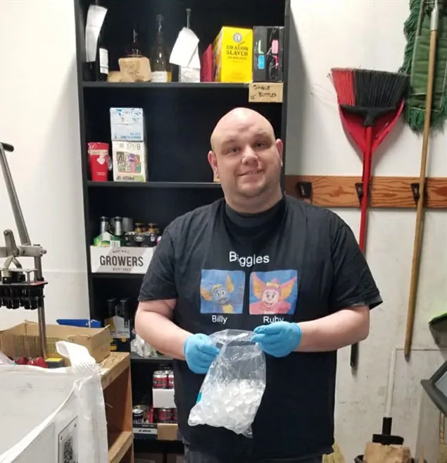 A man in a black t-shirt and blue gloves holding a bag of ice in a storage room
