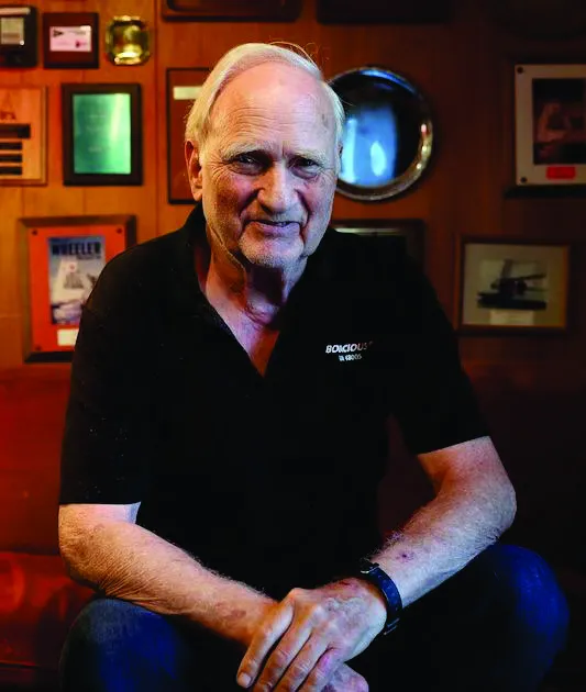 Portrait of John Clauser sitting in a room with a wood-paneled wall decorated with various framed awards and photographs