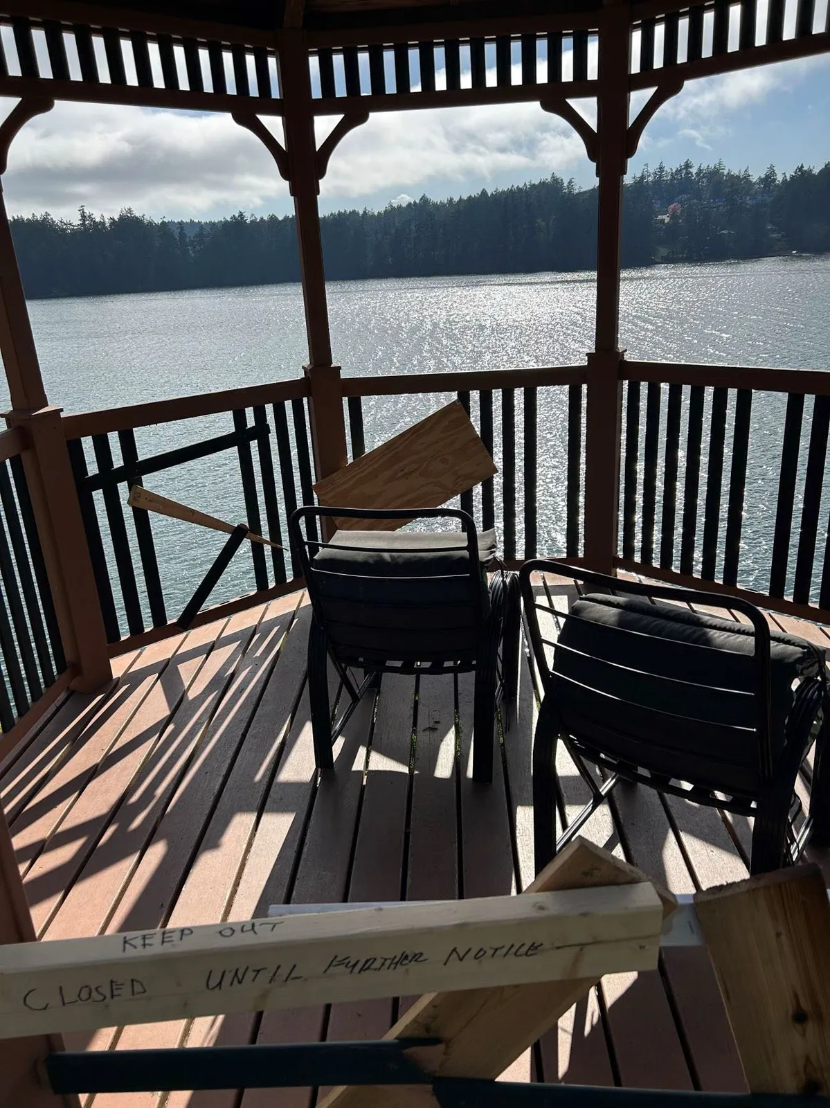 Interior view of a damaged wooden gazebo overlooking the water, with chairs inside and a temporary wooden barrier marked "CLOSED UNTIL FURTHER NOTICE"