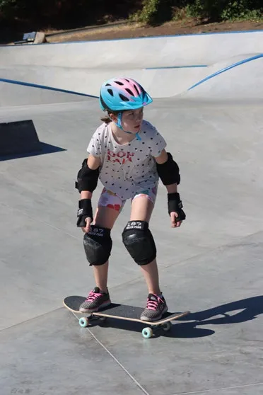 A young girl wearing a helmet and pads skateboarding at a skatepark