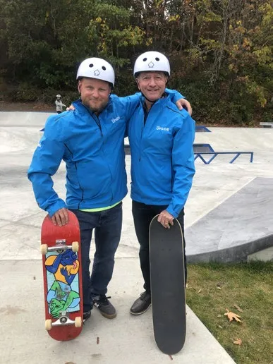 Two men in blue jackets wearing helmets and holding skateboards at a skatepark