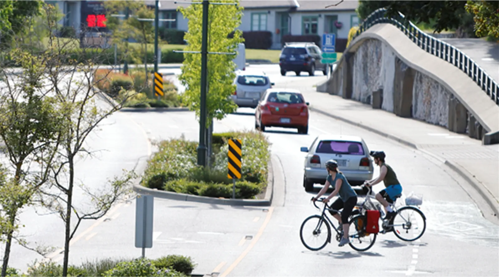 Two cyclists riding on a paved road crossing Island Highway, with vehicles in the background