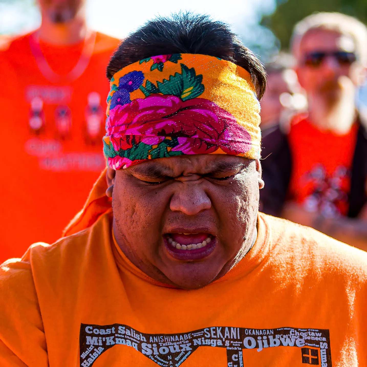 A man in an orange shirt and floral headband singing