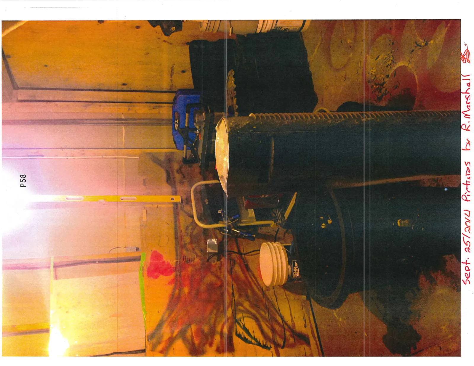 A room with buckets, equipment, and a large black cylindrical object next to a wall with red paint markings.