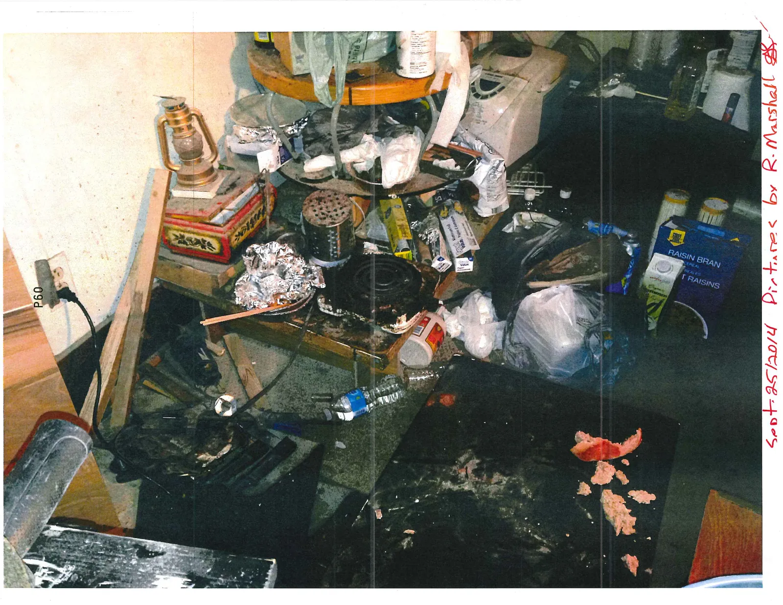 A cluttered area with a hot plate, a lantern, a box of Raisin Bran cereal, and other household items and trash.