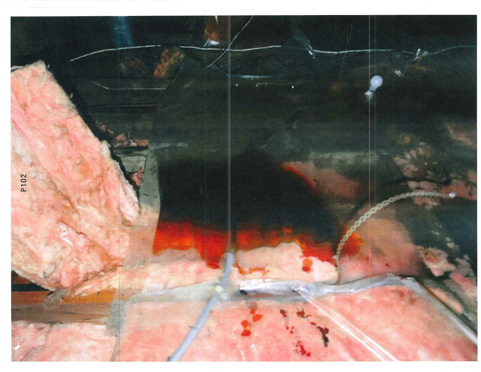 Photograph of an attic or crawlspace showing pink fiberglass insulation with significant dark reddish-brown staining. The image includes electrical wiring, a light bulb, and the label "P102" at the top.