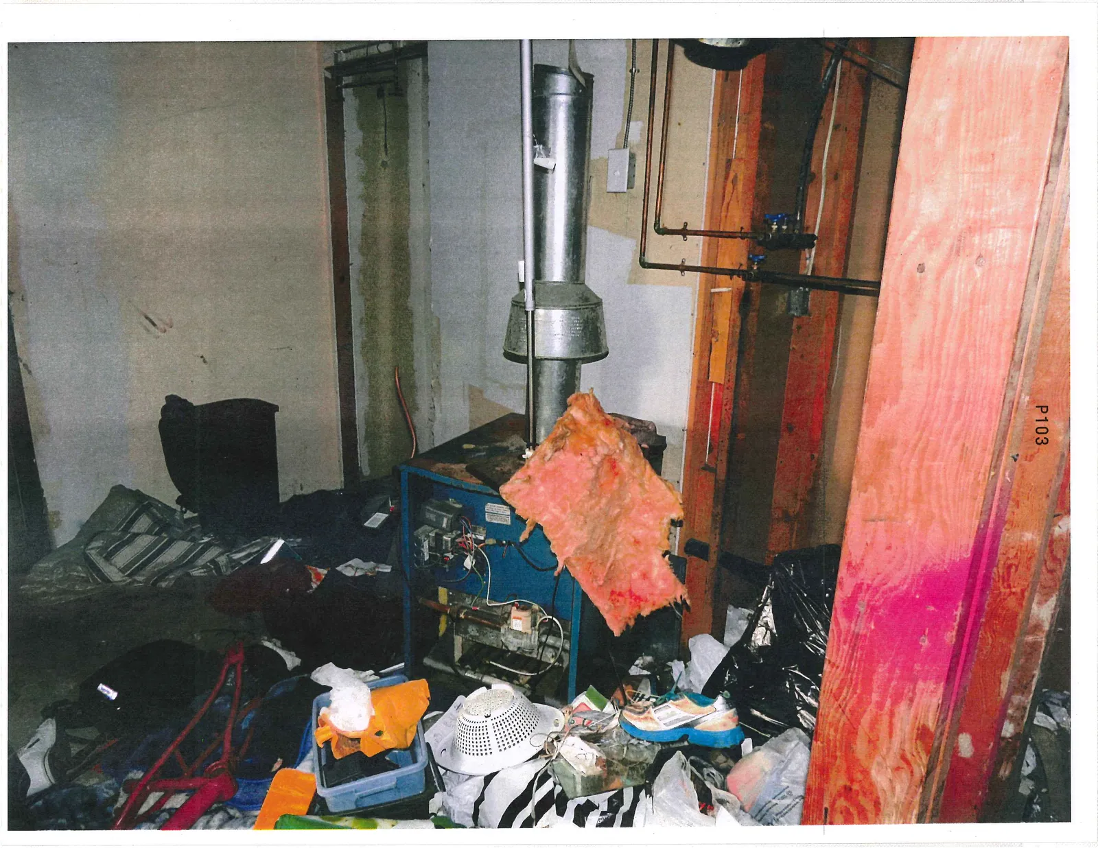 A photograph of a cluttered utility area featuring a blue furnace with a silver vent pipe. A piece of pink fiberglass insulation is draped over the vent pipe. The surrounding area is filled with various items including clothing, bags, a white colander, and storage bins. Copper piping and electrical conduit are visible on the wall in the background.