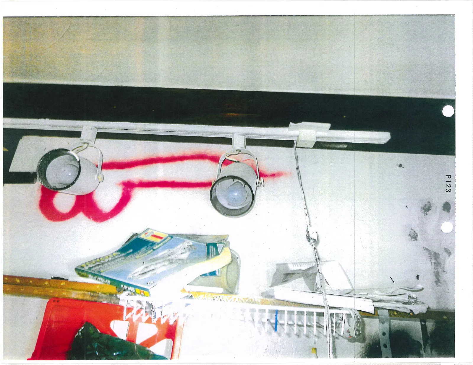 Track lighting fixture on a wall above red spray-painted graffiti and a wire shelf with magazines