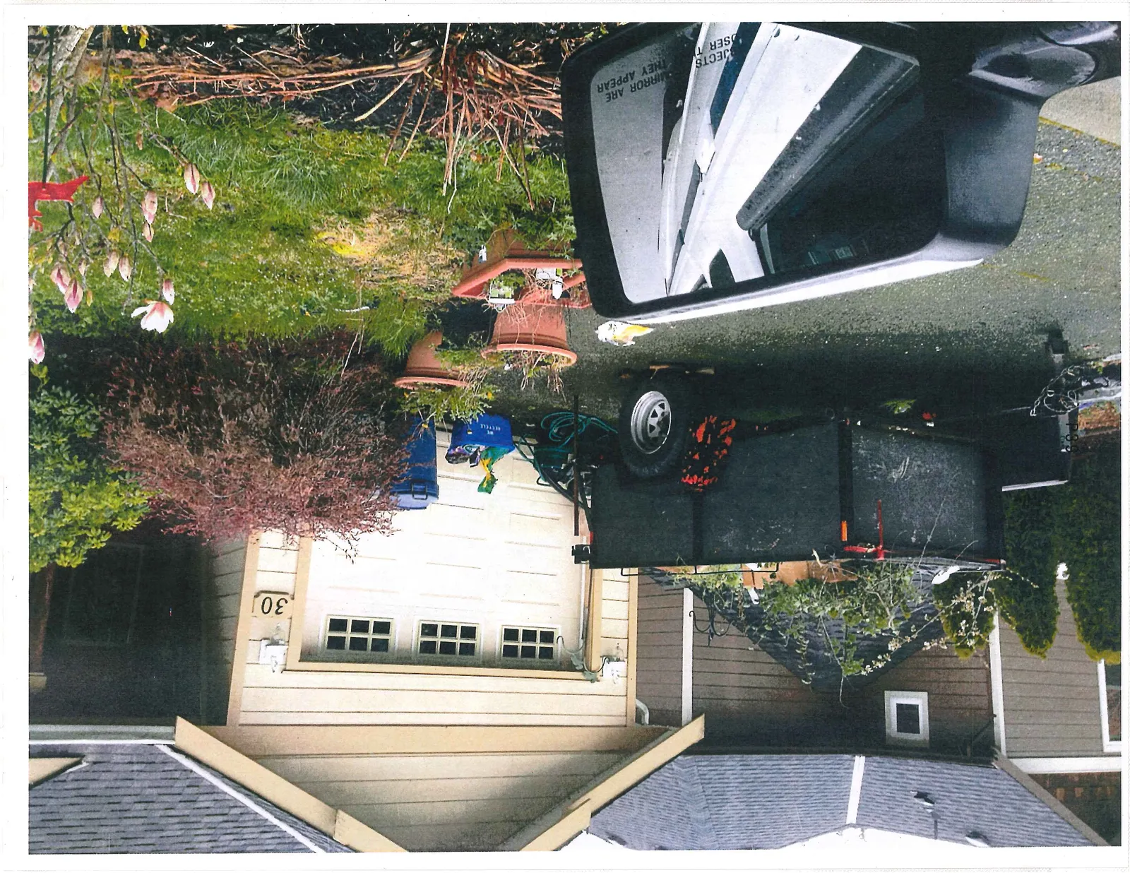 View of the driveway at address 30 reflected in a vehicle's side mirror, showing a utility trailer, potted plants, and recycling bins.