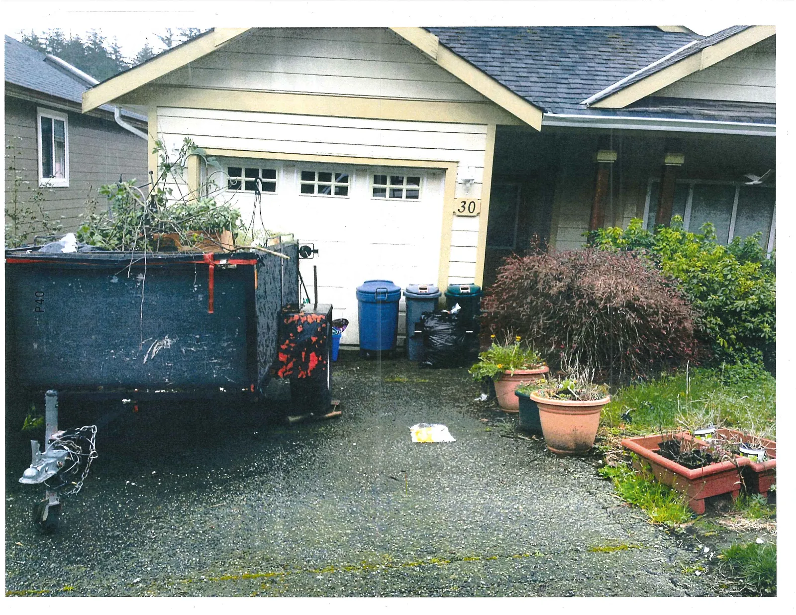 Perspective of the driveway at address 30, featuring a black utility trailer filled with yard debris, multiple waste and recycling containers, and several large potted plants.