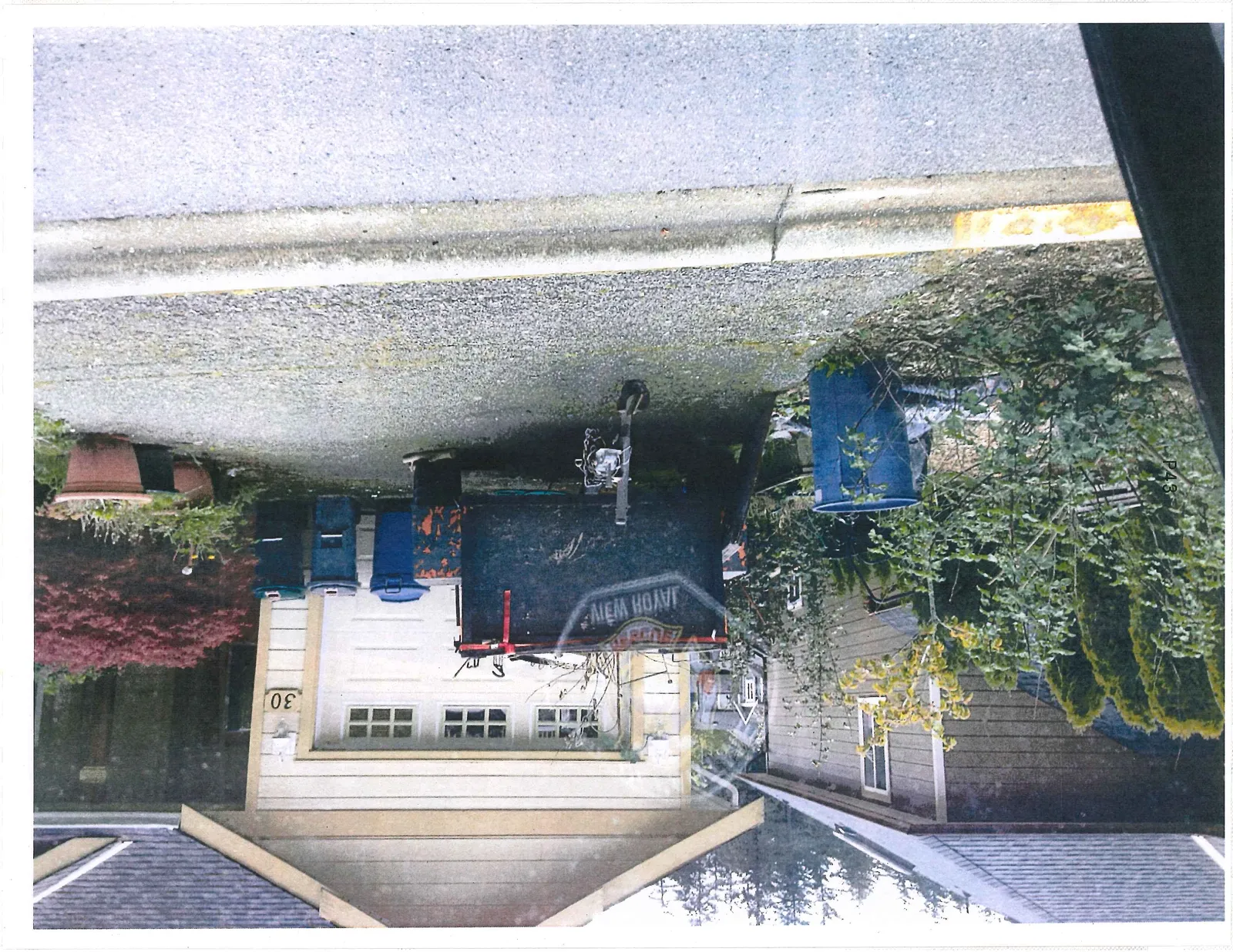 Exterior view of the driveway at house 30, featuring a black utility trailer with "VIEW ROYAL" text, blue recycling bins, and a red Japanese Maple bush.