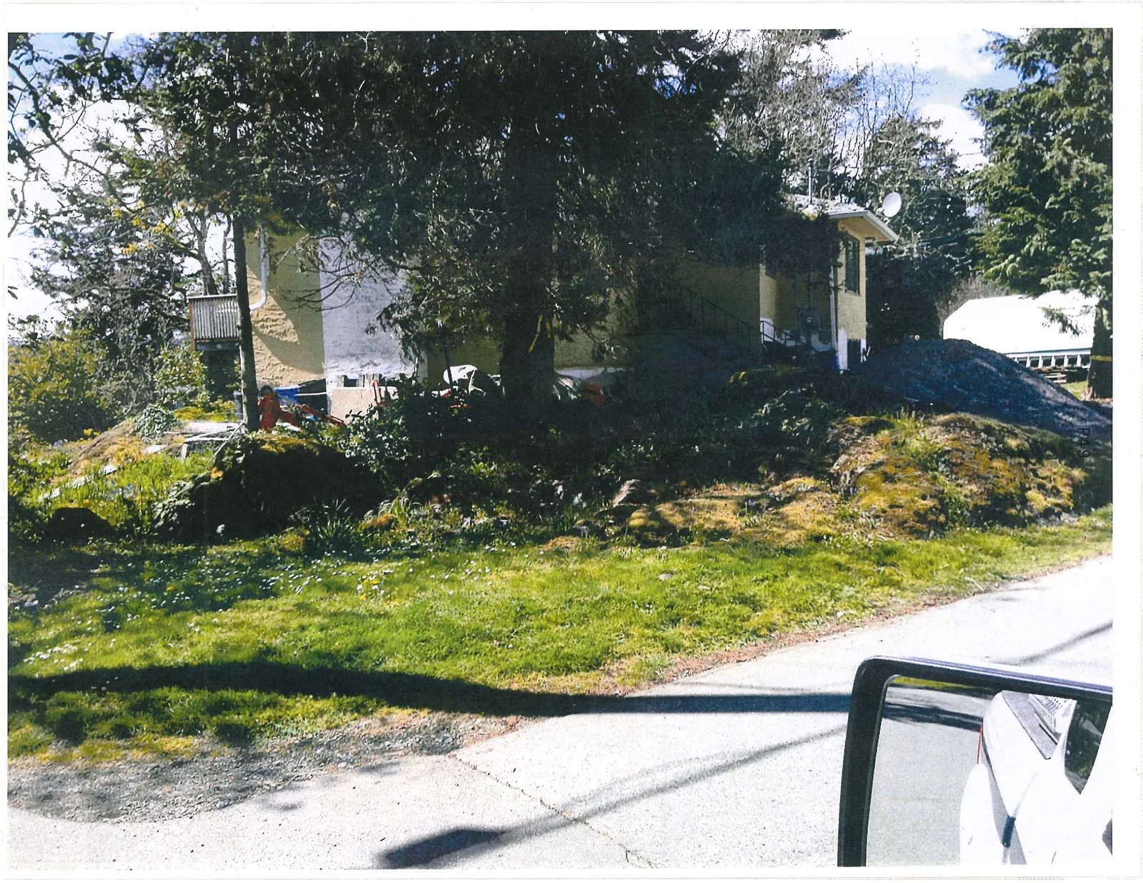 View of a two-story yellow house partially obscured by large trees, featuring a pile of gravel in the front yard and a vehicle's side-view mirror in the lower right foreground.