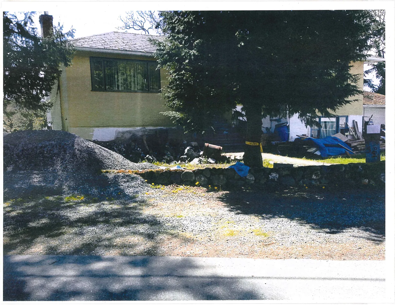 Front view of the yellow house showing a low stone wall bordering a gravel area, with various items and construction materials scattered in the yard.