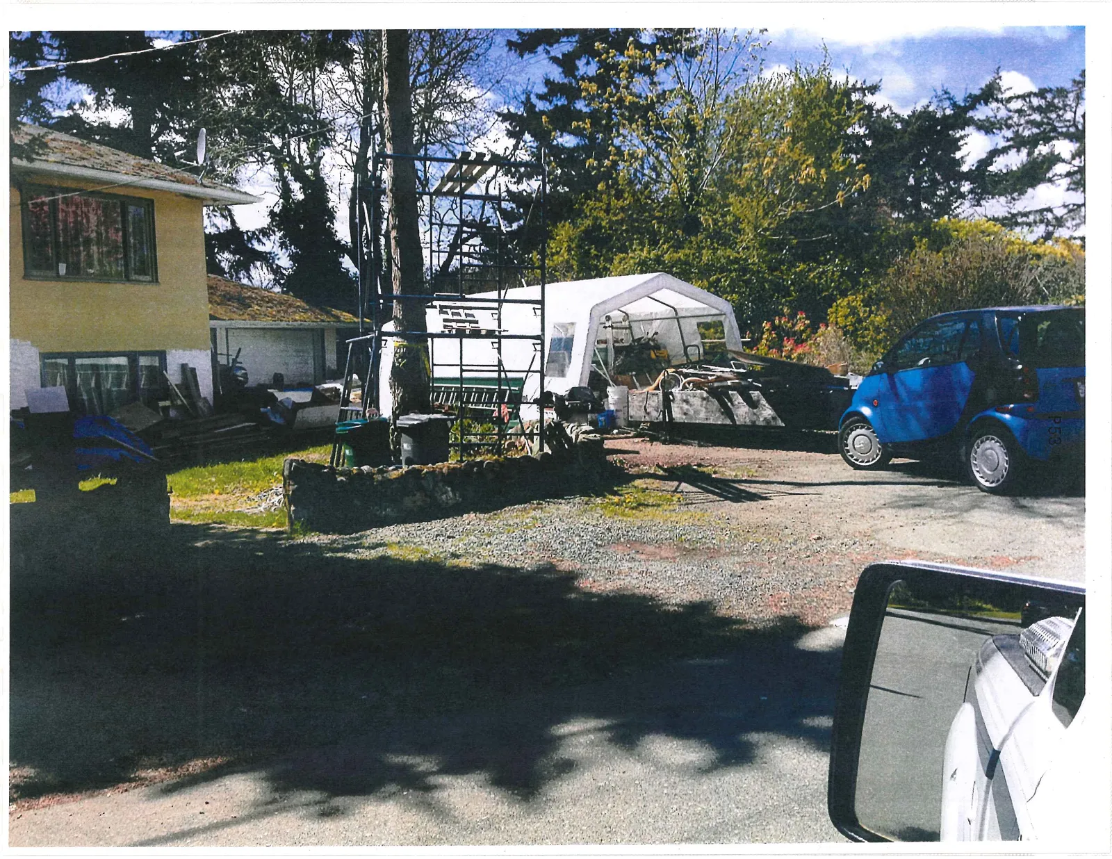 View of the driveway area showing a blue compact car parked near a white temporary structure and scaffolding, with a vehicle's side-view mirror visible in the upper right corner.