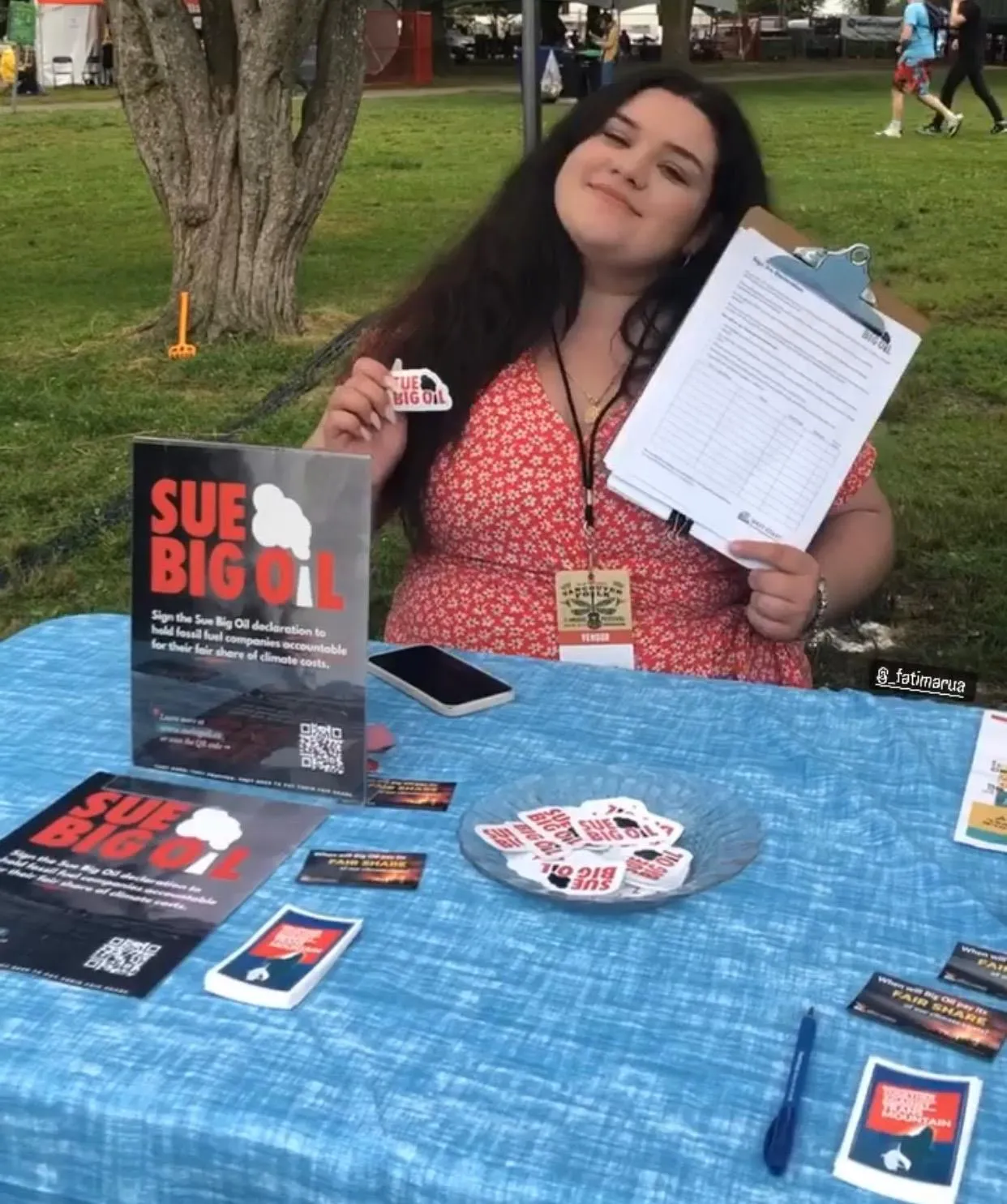 A woman standing at a campaign table for "Sue Big Oil" holding a clipboard and a sticker.