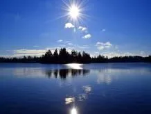 A landscape photo of a sun shining over a body of water with a treeline on the horizon.
