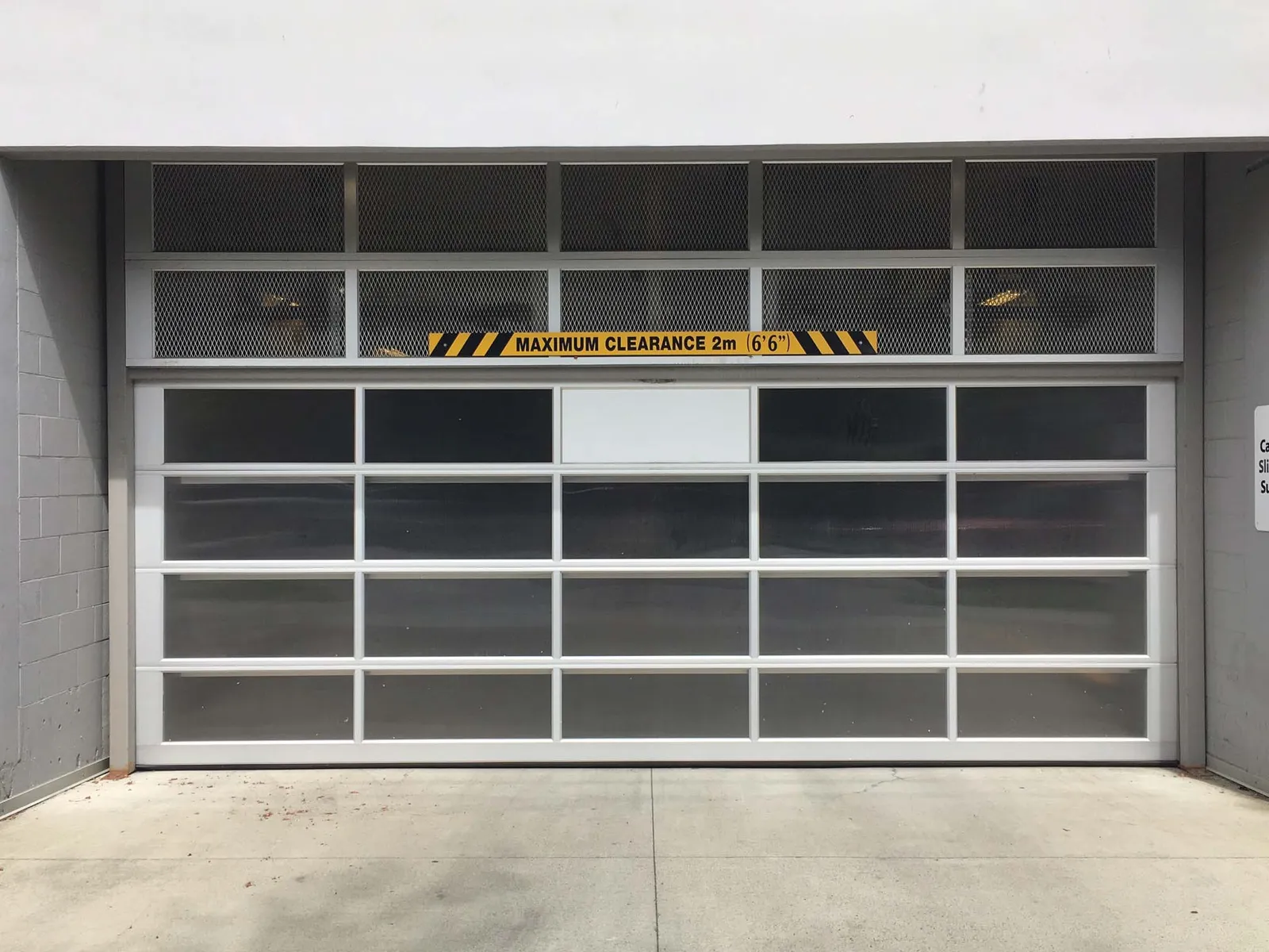 Photograph of an aluminum and polycarbonate panel overhead door featuring a yellow and black "MAXIMUM CLEARANCE 2m (6'6")" warning sign.