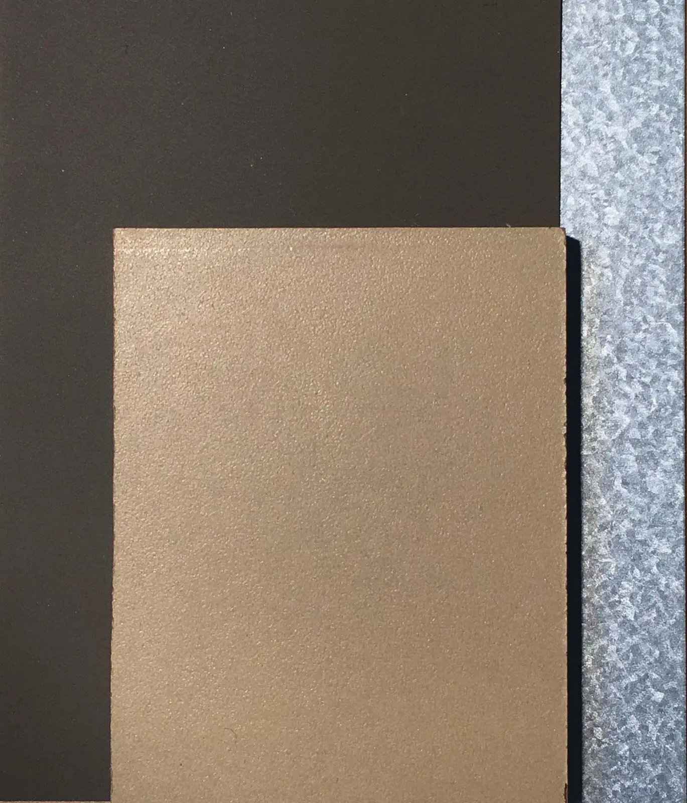 Photograph showing a collection of material samples, including dark metal flashing, painted metal, tan cement fibre board paneling, and a translucent polycarbonate panel.
