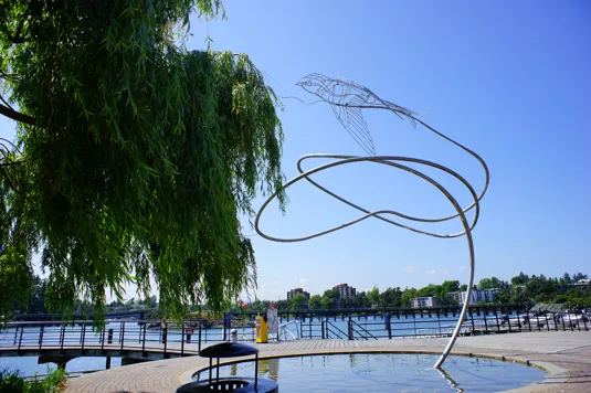 Metal bird sculpture near a willow tree by the water